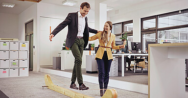 Ein Mann balanciert über eine Slackline im Büro. Eine Frau unterstützt ihn dabei. 