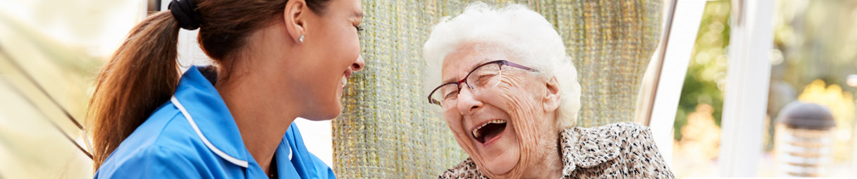 Junge Pflegerin sitzt bei Seniorin, beide lachen herzhaft.