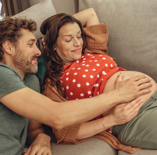 Schwanger Frau sitzt liegt auf dem Sofa. Neben ihr sitzt ihr Mann und beide haben ihre Hände auf dem Schwangerschaftsbauch.