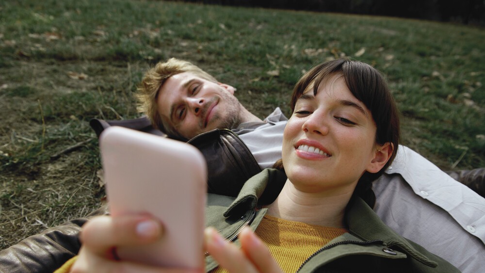 Ein Mann liegt im Gras. Auf seiner Brust liegt eine Frau, die ihr Smartphone in der Hand hält. 