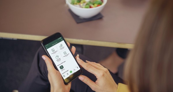 eine Frau sitzt am Tisch. Sie hat vor sich einen Salat stehen und sie schaut auf Ihr Smartphone, das die APP Meine AOK zeigt.