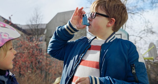 Ein Junge und ein Mädchen sind draußen und spielen. Der Junge steht mittig im Bild und benutzt gerade sein Asthma Spray. Das Mädchen steht mit ihrem Fahrradhelm auf dem Kopf am linken Bildrand.