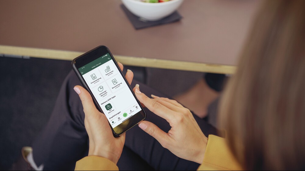 eine Frau sitzt am Tisch. Sie hat vor sich einen Salat stehen und sie schaut auf Ihr Smartphone, das die APP Meine AOK zeigt.