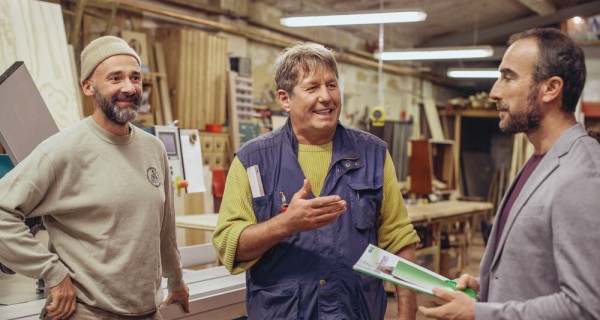 Drei Männer stehen in einer Werkstatt zusammen. Der rechte Mann ist ein AOK-Berater. Die beiden anderen Männer links hören ihm freundlich zu.