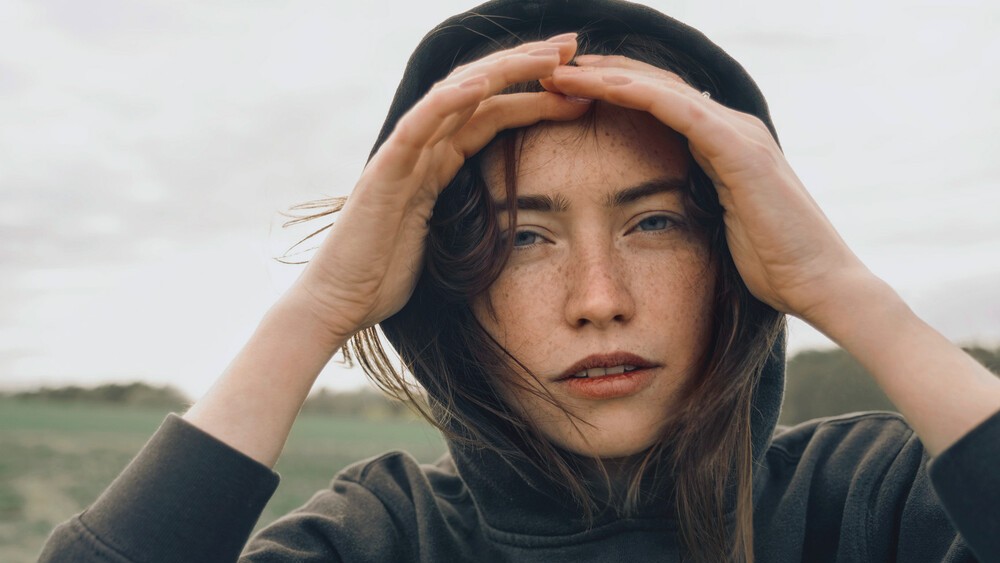 Eine junge Frau steht in der Natur und schaut mit ihren Händen in die Ferne. Sie trägt eine Kapuze. 