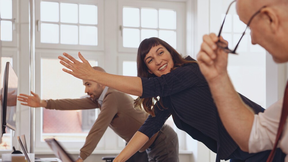 In einem Büro sind zwei Männer und eine Frau. Sie haben alle einen PC oder Laptop vor sich. Rechts außen ist ein Mann, der gerade seine Brille absetzt und zu den anderen Kollegen schaut. Hinter ihm steht eine Frau, die ihn freundlich anlacht. Sie und der dritte Kollege hinter ihr stehen nach vorne gebeugt und halten jeweils einen Arm nach oben zur Mobilisation. 