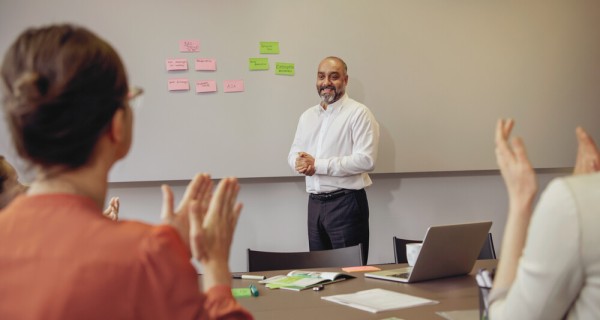 Blick in einen Schulungsraum von hinten. Zwei Personen sitzen links und rechts und klatschen. Der Präsentator steht vorne und hat auf der Wand Pinnkarten neben sich. 