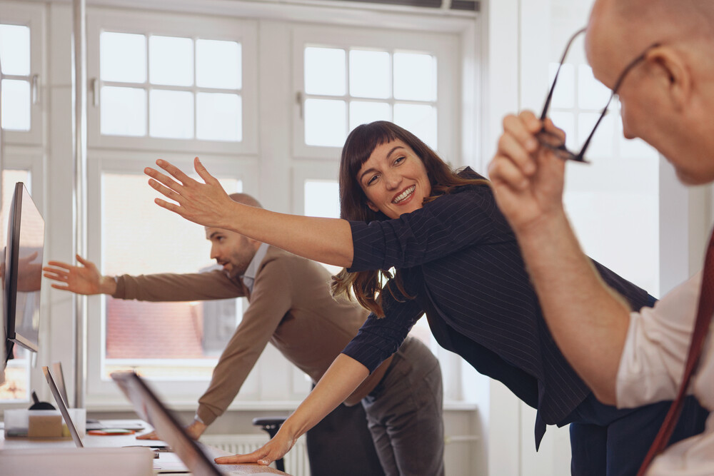 In einem Büro sind zwei Männer und eine Frau. Sie haben alle einen PC oder Laptop vor sich. Rechts außen ist ein Mann, der gerade seine Brille absetzt und zu den anderen Kollegen schaut. Hinter ihm steht eine Frau, die ihn freundlich anlacht. Sie und der dritte Kollege hinter ihr stehen nach vorne gebeugt und halten jeweils einen Arm nach oben zur Mobilisation.