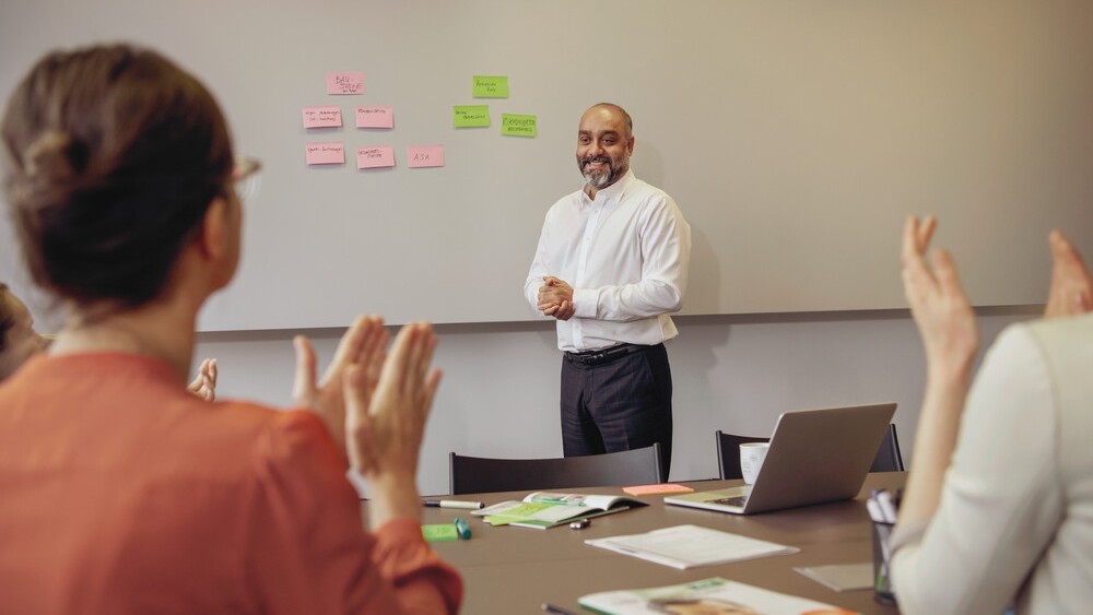 Blick in einen Schulungsraum von hinten. Zwei Personen sitzen links und rechts und klatschen. Der Präsentator steht vorne und hat auf der Wand Pinnkarten neben sich. 