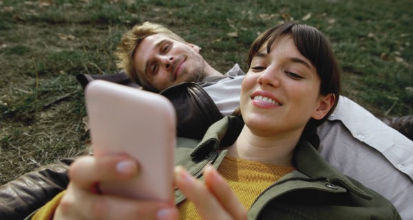 Ein Mann liegt im Gras. Auf seiner Brust liegt eine Frau, die ihr Smartphone in der Hand hält. 