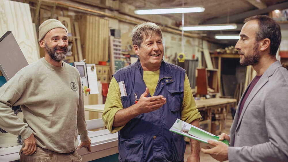 Drei Männer stehen in einer Werkstatt zusammen. Der rechte Mann ist ein AOK-Berater. Die beiden anderen Männer links hören ihm freundlich zu.