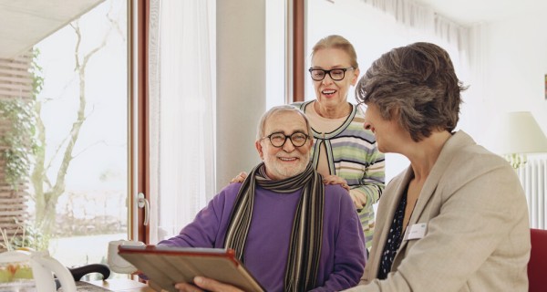 AOK-Mitarbeiterin schaut lächelnd den neben ihr sitzenden Mann an. Der Mann lächelt zurück. Hinter ihm steht eine Frau, die ihre Hände auf seine Schultern legt.