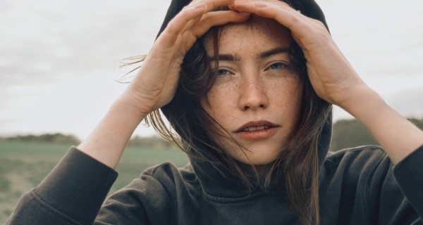 Eine junge Frau steht in der Natur und schaut mit ihren Händen in die Ferne. Sie trägt eine Kapuze. 
