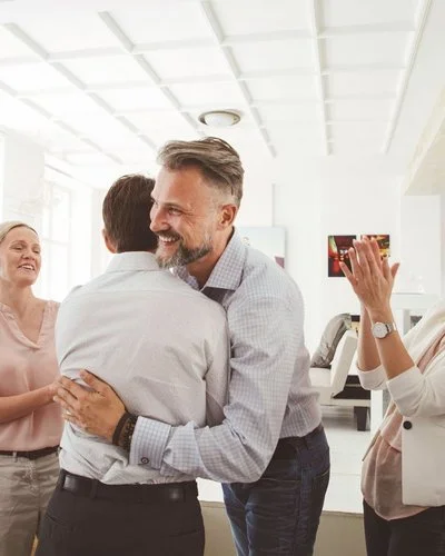 Zwei Kollegen umarmen sich im Büro, drei Menschen stehen um sie herum und applaudieren