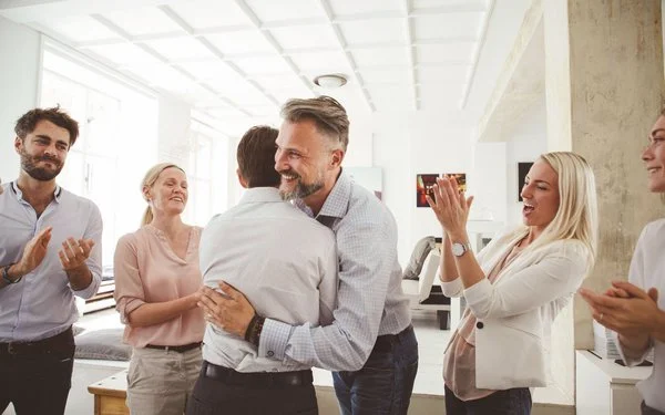 Zwei Kollegen umarmen sich im Büro, drei Menschen stehen um sie herum und applaudieren