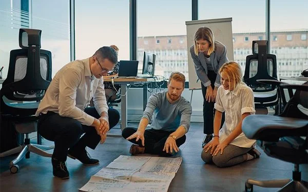 Vier Beschäftigte sitzen und knien gemeinsam auf dem Büroboden und besprechen Ergebnisse eines Workshops zur Stressreduktion.