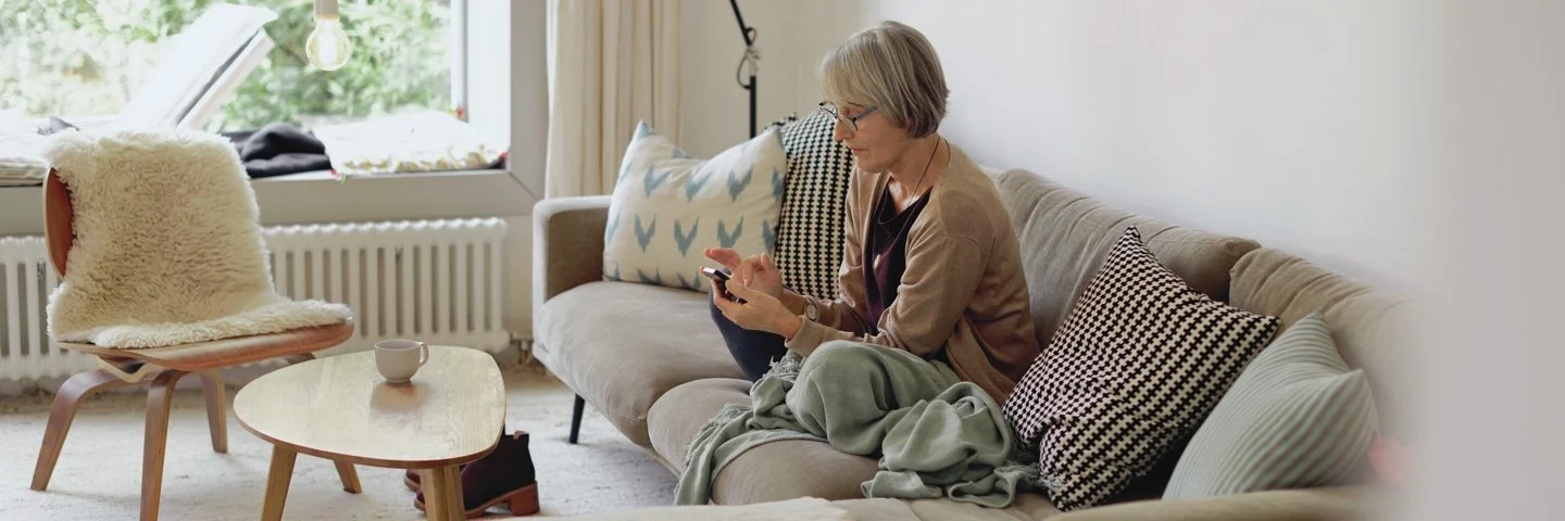 Eine ältere Frau sitzt auf einem Sofa und nutzt ein Smartphone.
