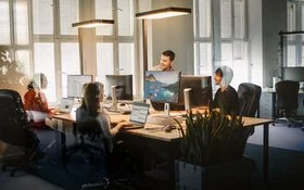 Vier Personen sitzen im Großraumbüro an zusammengestellten Tischen vor ihren Laptops und Bildschirmen und unterhalten sich