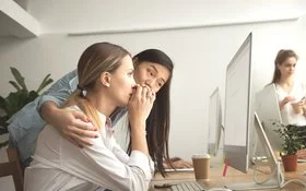Eine junge Frau sitzt am Schreibtisch und schaut beunruhigt auf ihren Laptop, eine Kollegin legt den Arm um sie