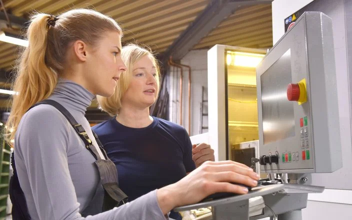 In einem Produktionsbetrieb. Eine Kollegin erläutert einer anderen etwas an einem Monitor.