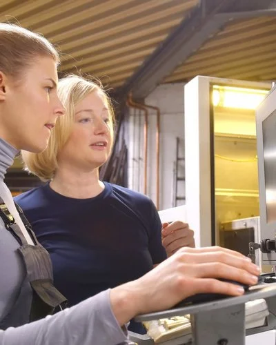 In einem Produktionsbetrieb. Eine Kollegin erläutert einer anderen etwas an einem Monitor.