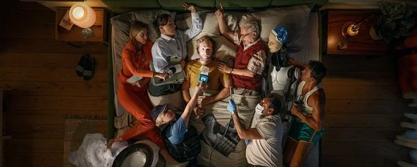 Guter Schlaf zahlt sich aus: Ein Mann liegt wach im Bett, von oben fotografiert. Um ihn drängen sich Figuren, die für seine Gedanken stehen, darunter eine Reporterin mit Mikrofon, ein Arzt, eine Kollegin mit Akten, ein Roboter und ein Sportler.