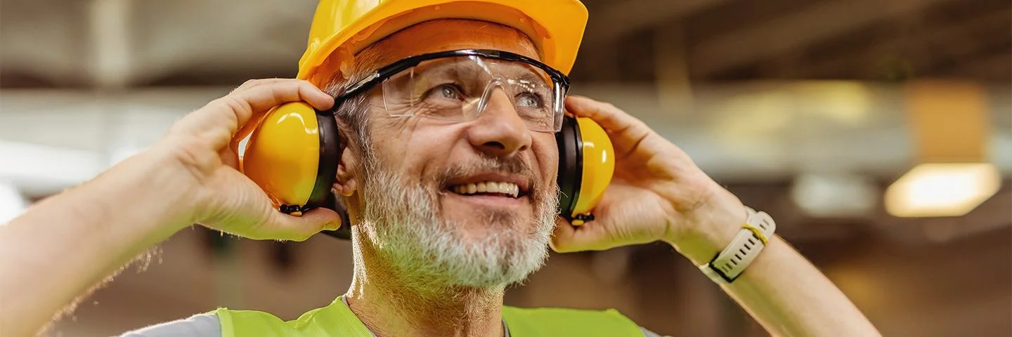 Ein lächelnder Bauarbeiter mit Helm hebt mit beiden Händen seine Ohrenschützer von den Ohren und blickt nach oben.