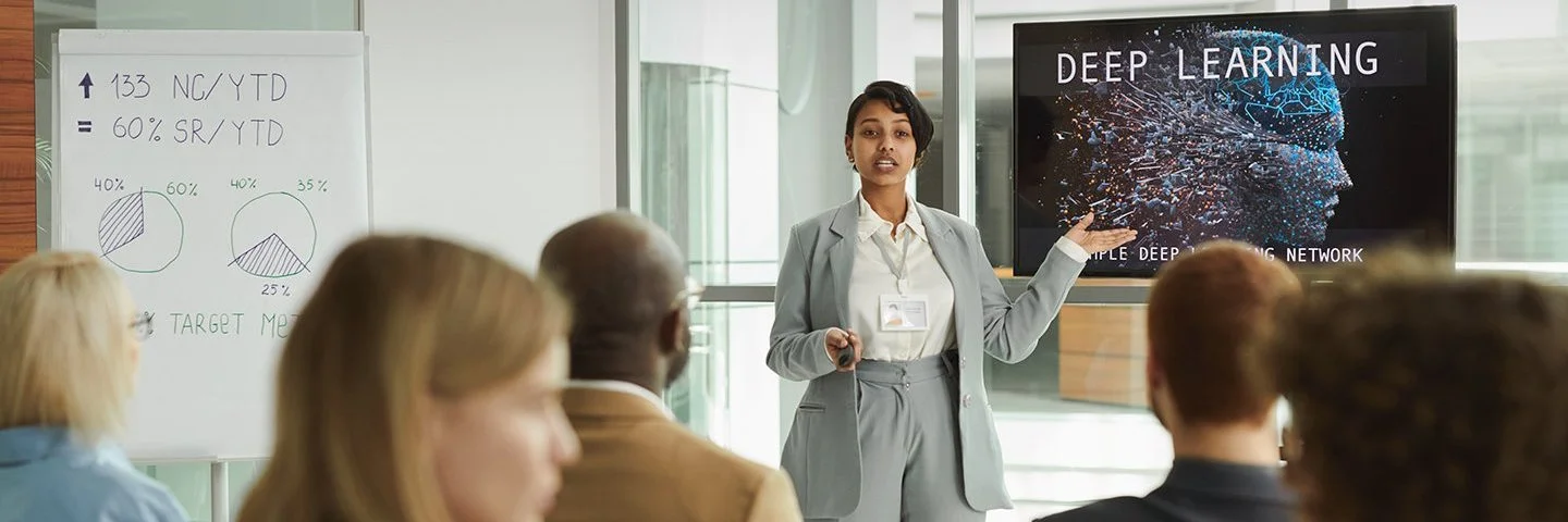 Eine Frau hält ein Seminar und erklärt den Teilnehmenden Inhalte an einer digitalen Tafel.