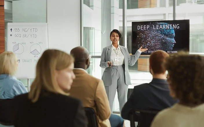 Eine Frau hält ein Seminar und erklärt den Teilnehmenden Inhalte an einer digitalen Tafel.