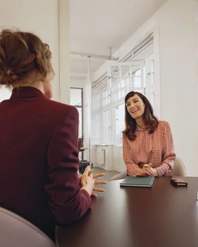 Eine Frau sitzt einer anderen Frau gegenüber an einem Konferenztisch in einem tageslichthelles Büro.