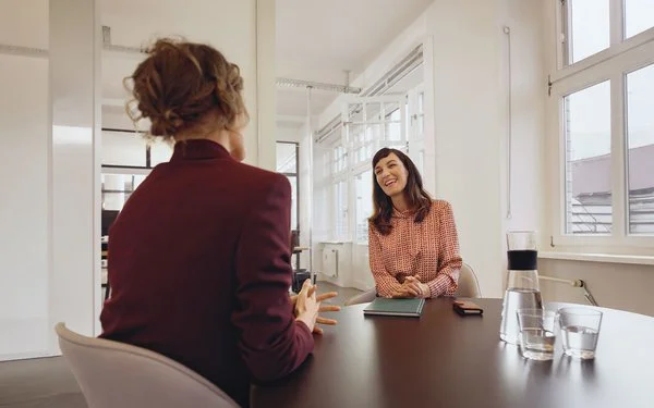 Eine Frau sitzt einer anderen Frau gegenüber an einem Konferenztisch in einem tageslichthelles Büro.