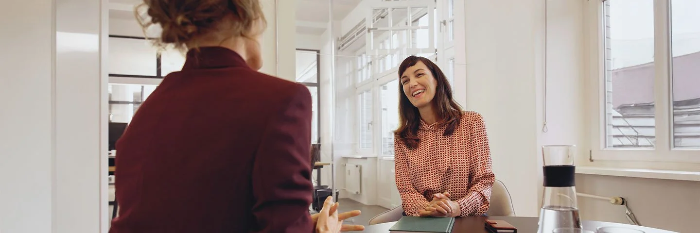Eine Frau sitzt einer anderen Frau gegenüber an einem Konferenztisch in einem tageslichthelles Büro.
