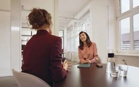 Eine Frau sitzt einer anderen Frau gegenüber an einem Konferenztisch in einem tageslichthelles Büro.