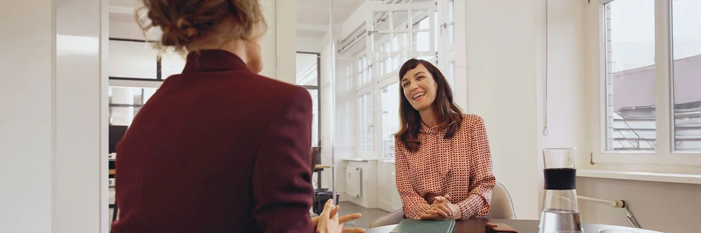 Eine Frau sitzt einer anderen Frau gegenüber an einem Konferenztisch in einem tageslichthelles Büro.