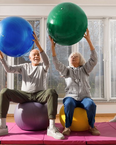 Pflegekraft leitet zwei ältere Personen zu Bewegungsübungen mit Petzibällen an.