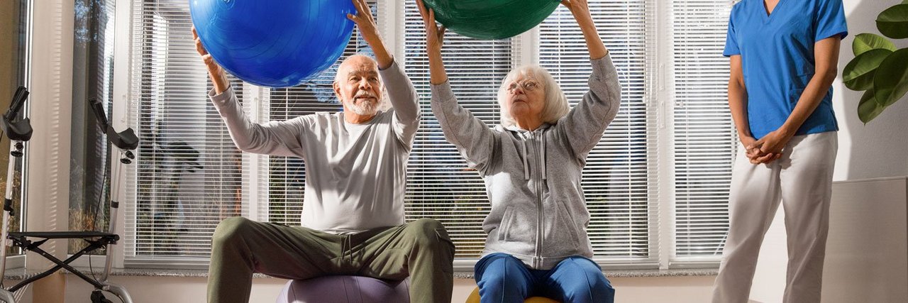 Pflegekraft leitet zwei ältere Personen zu Bewegungsübungen mit Petzibällen an.