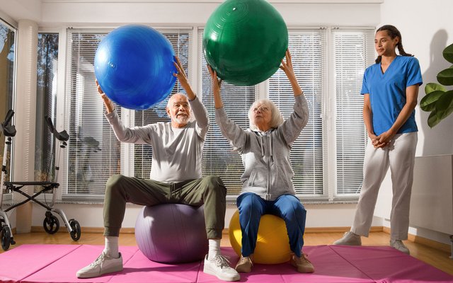 Pflegekraft leitet zwei ältere Personen zu Bewegungsübungen mit Petzibällen an.