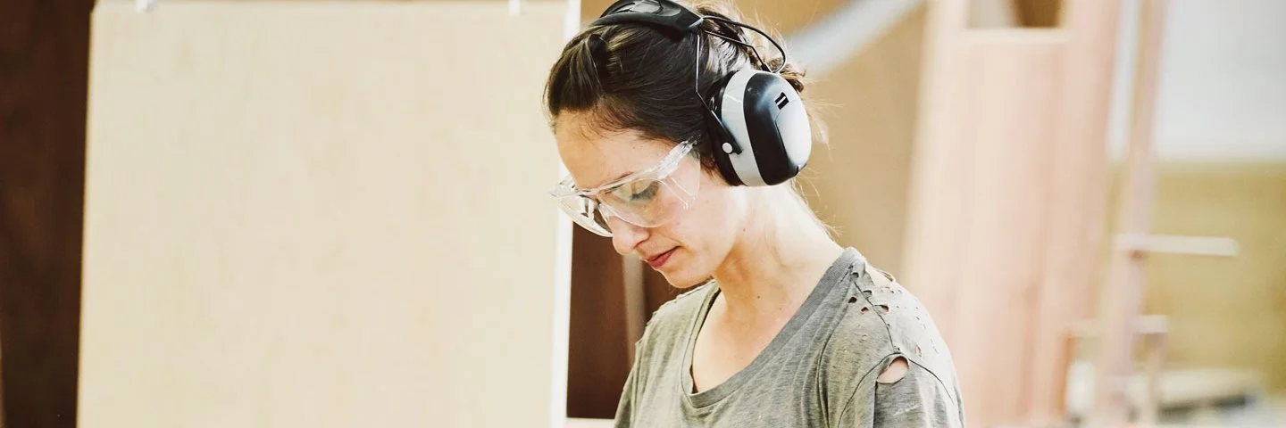 Eine Frau mit Gehörschutz und Schutzbrille steht in einer Werkstatt