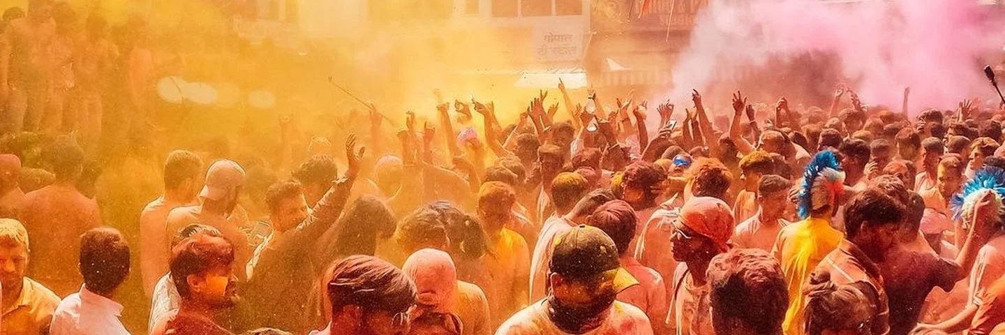 Eine Menschenmenge feiert das Holi-Festival mit buntem Farbnebel in Gelb und Pink, die Feiernden strecken ihre Arme in die Luft