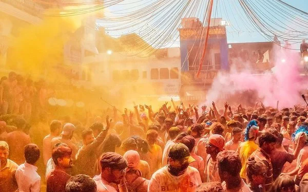 Eine Menschenmenge feiert das Holi-Festival mit buntem Farbnebel in Gelb und Pink, die Feiernden strecken ihre Arme in die Luft