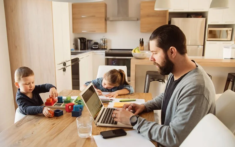 Ein Mann arbeitet am Laptop in der Küche, neben ihm ein Mädchen mit Spielsachen.