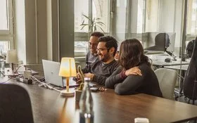 Drei Mitarbeitende sitzen gut gelaunt nebeneinander an einem Tisch und schauen gemeinsam auf einen geöffneten Laptop.