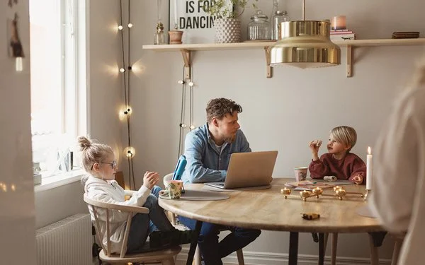 Ein Mann arbeitet zu Hause am Laptop, während seine zwei Kinder neben ihm am Tisch sitzen.