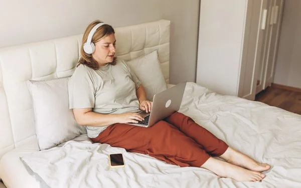 Eine Frau sitzt mit Laptop und Kopfhörern auf ihrem Bett