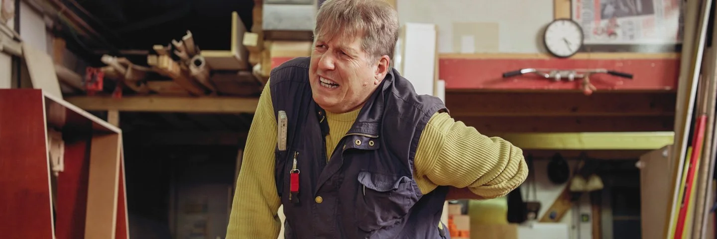 Ein Mann mit schmerzverzerrtem Gesicht steht in einer Werkstatt und stütz seinen Rücken.