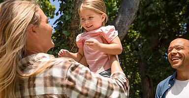 Familienfreundlichkeit in Unternehmen: Eine Frau hebt ein Kind im Park hoch, während ein Mann danebensteht.