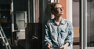 Gesunde Pause: Junge Frau mit Brille und Jeanshemd lehnt entspannt an einem Fenster im Sonnenlicht und hält ein Notizbuch in den Händen Gesunde Pause: Junge Frau mit Brille und Jeanshemd lehnt entspannt an einem Fenster im Sonnenlicht und hält ein Notizbuch in den Händen