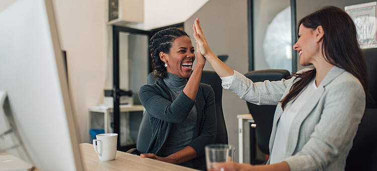 Zwei Frauen klatschen „Highfive“ am Schreibtisch sitzend