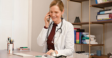 Telefonische-AU Zu sehen ist eine Ärztin, die an einem Schreibtisch sitzt und telefoniert.