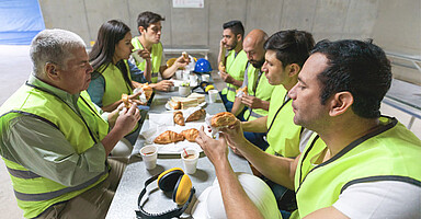 Eine Gruppe von Frauen und Männern in Arbeitskleidung sitzt an einem großen Tisch. Sie essen und trinken. 
